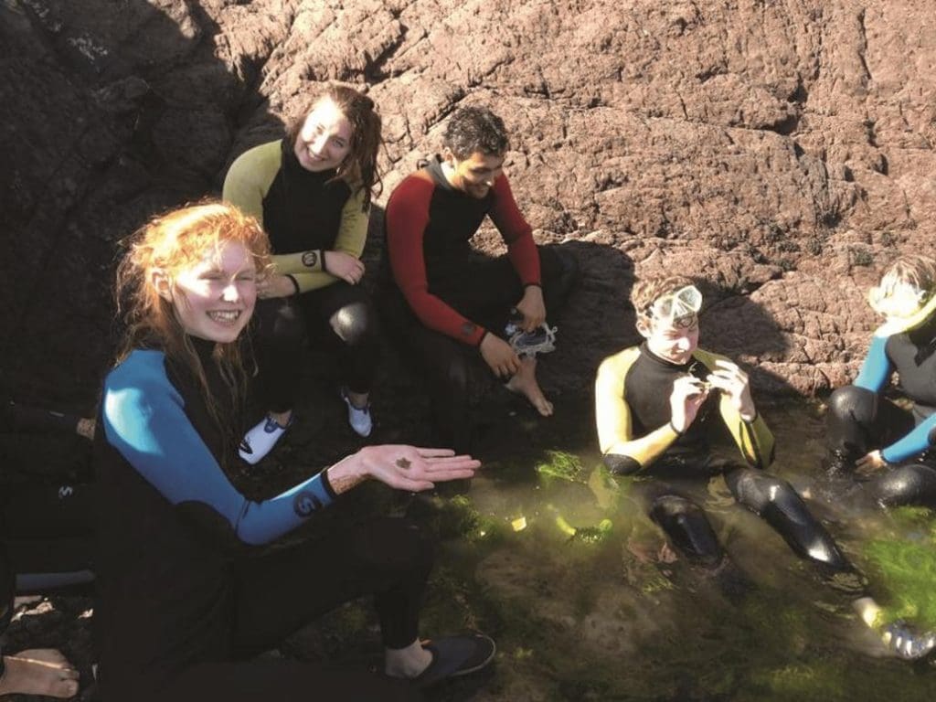 school group in wets suits sitting in a rock pool at the seaside