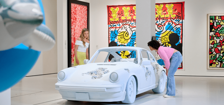 a white sculpture of a sports car in an exhibition room with students looking at it