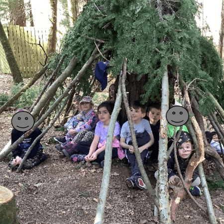 primary school pupils sitting under a build den in the woods made of logs and fern branches