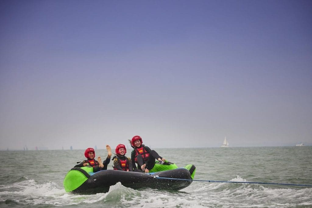 three school pupils in wet gear on a green inflatable on the sea being towed along by speed boat